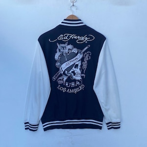May include: A black and white varsity jacket with white sleeves. The back of the jacket features a large Ed Hardy logo with a skull and crossbones design. The text "Ed Hardy", "USA", and "Los Angeles" is printed on the jacket.