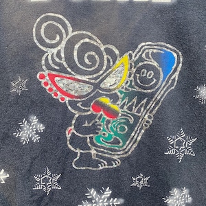 K&ouml;nnte beinhalten: Dunkelmarineblaues Sweatshirt mit einem skurrilen Cartoon-Charakter-Design. Die Figur hat silberne Umrisse, rote, gelbe und blaue Akzente und h&auml;lt ein farbenfrohes Objekt. Silberne Schneeflocken sind um das Design verstreut.