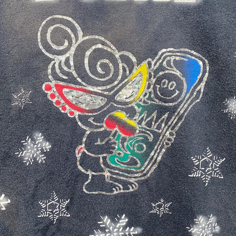 K&ouml;nnte beinhalten: Dunkelmarineblaues Sweatshirt mit einem skurrilen Cartoon-Charakter-Design. Die Figur hat silberne Umrisse, rote, gelbe und blaue Akzente und h&auml;lt ein farbenfrohes Objekt. Silberne Schneeflocken sind um das Design verstreut.