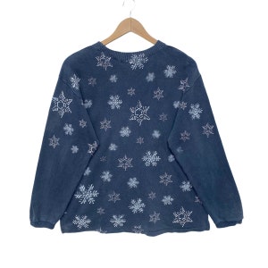 K&ouml;nnte beinhalten: Ein dunkelblaues Sweatshirt mit einem Muster aus wei&szlig;en Schneeflocken. Das lang&auml;rmlige Oberteil h&auml;ngt an einem Holzb&uuml;gel. Die Schneeflocken sind unterschiedlich gro&szlig; und gleichm&auml;&szlig;ig &uuml;ber den Stoff verteilt.