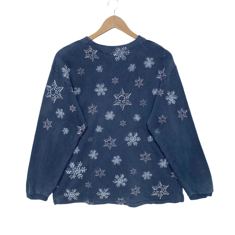 K&ouml;nnte beinhalten: Ein dunkelblaues Sweatshirt mit einem Muster aus wei&szlig;en Schneeflocken. Das lang&auml;rmlige Oberteil h&auml;ngt an einem Holzb&uuml;gel. Die Schneeflocken sind unterschiedlich gro&szlig; und gleichm&auml;&szlig;ig &uuml;ber den Stoff verteilt.