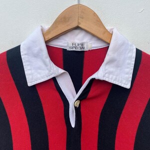 May include: A red, black, and white striped rugby shirt with a white collar. The shirt has the text "FLIPS SPECIAL" on the label. The shirt is hanging on a wooden hanger.