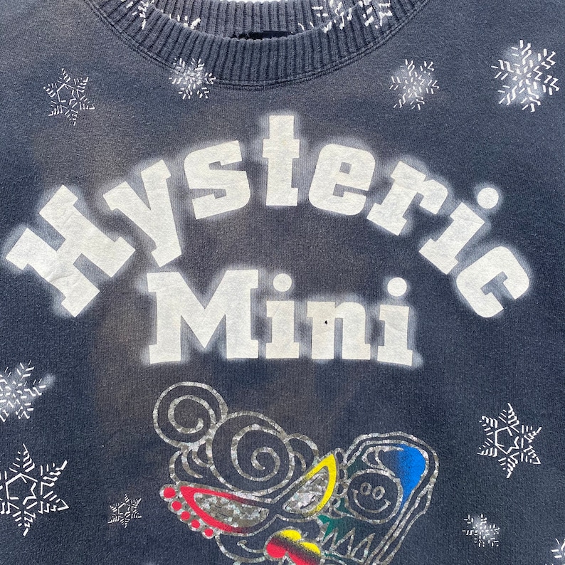 K&ouml;nnte beinhalten: Dunkelblaues Sweatshirt mit dem wei&szlig;en Schriftzug "Hysteric Mini" in einem Bogen auf der Vorderseite. Das Design umfasst eine Cartoonfigur mit farbenfrohen Akzenten und Schneeflocken. Kragen und Manschetten sind gerippt.