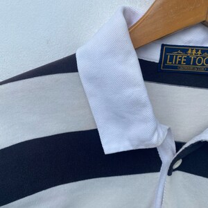 May include: A white and navy blue striped polo shirt with a white collar. The shirt has a logo patch on the left chest that reads "Life Too Short" with a small mountain range graphic above the text.