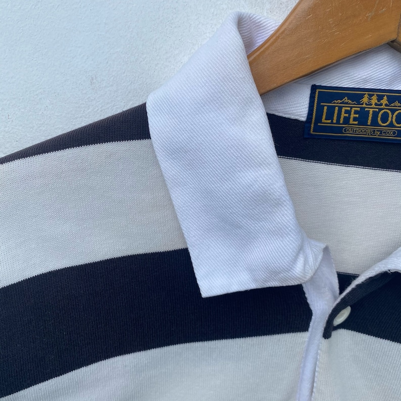 May include: A white and navy blue striped polo shirt with a white collar. The shirt has a logo patch on the left chest that reads "Life Too Short" with a small mountain range graphic above the text.