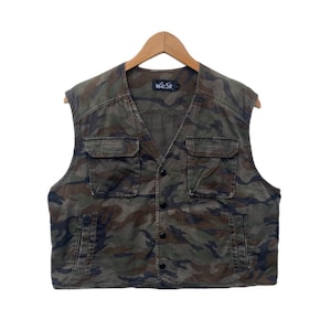 Peut inclure: Gilet de camouflage avec quatre poches avant et une fermeture boutonnée. Le gilet est dans les tons de vert, marron et noir. La marque "W&SB" est visible en haut du dos. Le gilet est suspendu à un cintre en bois.