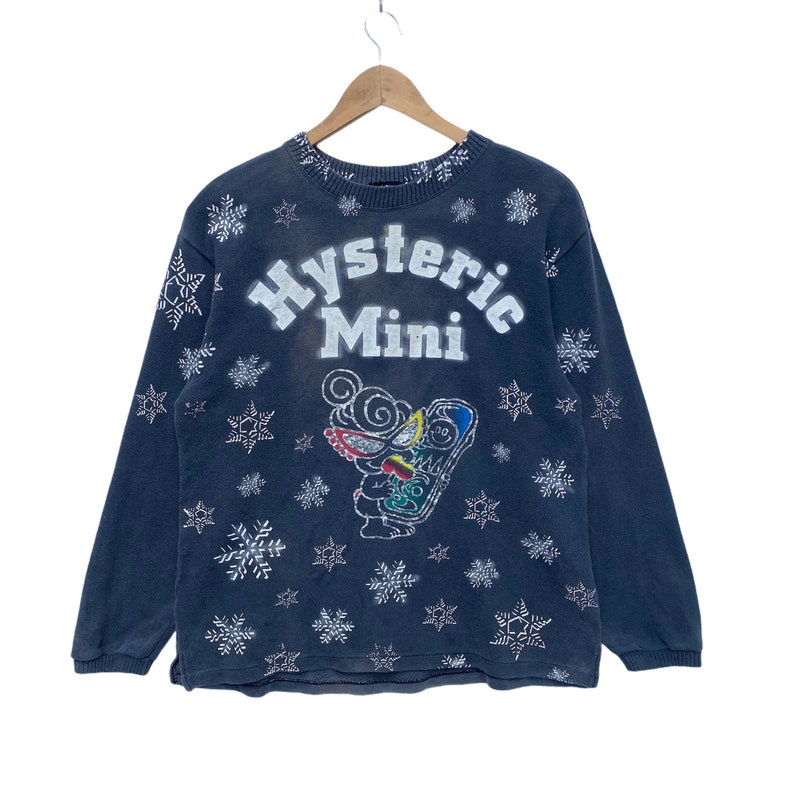 K&ouml;nnte beinhalten: Dunkelblaues Sweatshirt mit dem Text "Hysteric Mini" in Wei&szlig;, mit einer Cartoon-Grafik und Schneeflockenmustern. Das lang&auml;rmlige Oberteil h&auml;ngt an einem Holzkleiderb&uuml;gel.