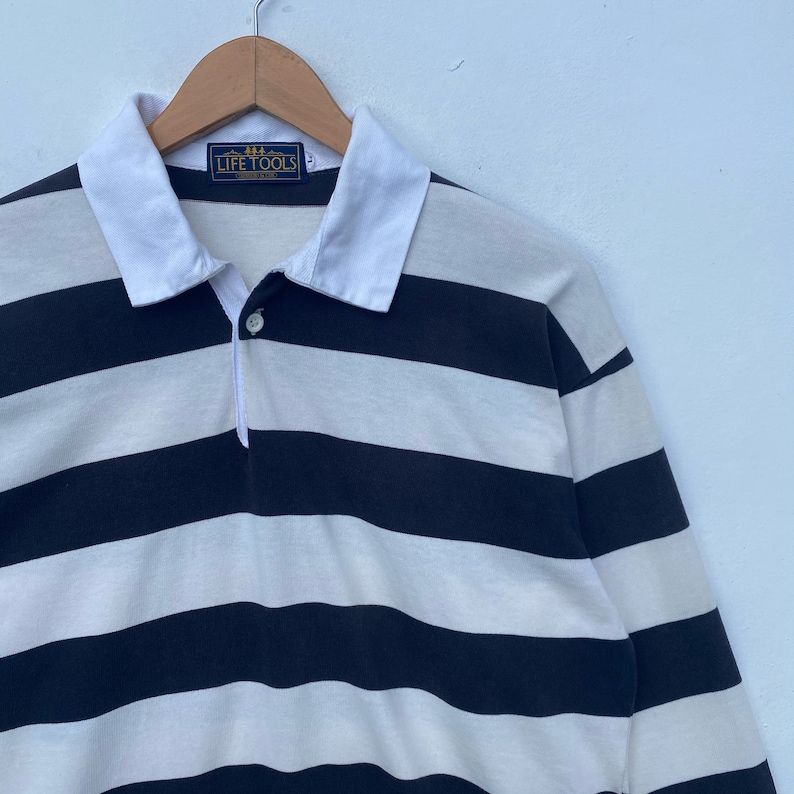 May include: A long-sleeved striped polo shirt with a white collar and a button closure. The shirt is black and white with horizontal stripes. The shirt has a label that reads "Life Tools" on the front.
