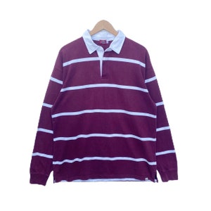 Puede incluir: Una camiseta de rugby de manga larga con rayas blancas sobre un fondo burdeos. La camiseta tiene un cuello abotonado y una tapeta de dos botones.