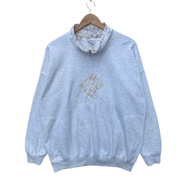 May include: A gray sweatshirt with a floral print on the inside of the collar. The sweatshirt has a half-zip closure and a diamond-shaped logo with the text "Monica Boutique" embroidered on the front.