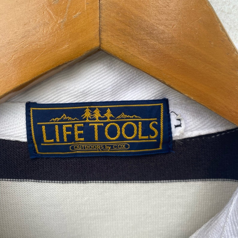 May include: A blue and gold label with the text "LIFE TOOLS OUTDOORS by COX" and a graphic of three pine trees and mountains.