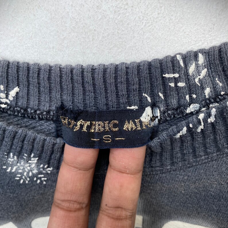 K&ouml;nnte beinhalten: Nahaufnahme eines dunkelgrauen Sweatshirts mit wei&szlig;en Schneeflockenmustern. Das Etikett lautet "HYSTERIC MINI" in goldfarbenen Buchstaben, darunter die Gr&ouml;&szlig;e "S". Der Stoff hat eine gerippte Textur.