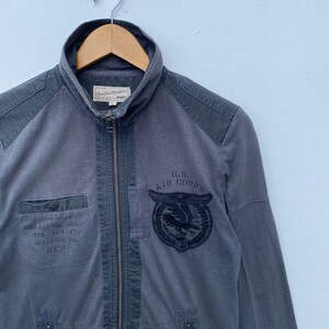 May include: A gray zip-up jacket with a U.S. Air Corps patch on the left chest. The jacket has a  "7500 ART. SIG. USAAF 420 ACT. A10 ALLIANCE AA. N&deg;19" text on the left side.