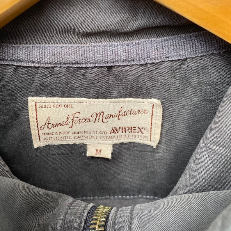 May include: A gray cotton jacket with a zipper closure. The label reads "Good for One Armed Forces Manufacturer AVIREX Name & Trade Mark Registered Authentic Garment Established in 1975" and has a size M tag.