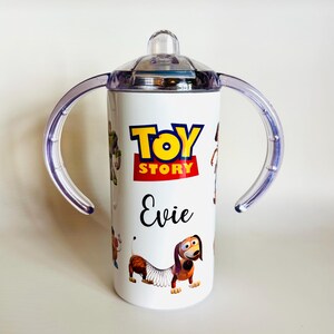 May include: White sippy cup with the "Toy Story" logo in yellow and red, and the name "Evie" in black script. The cup has a clear lid, clear handles, and illustrations of characters from the film.