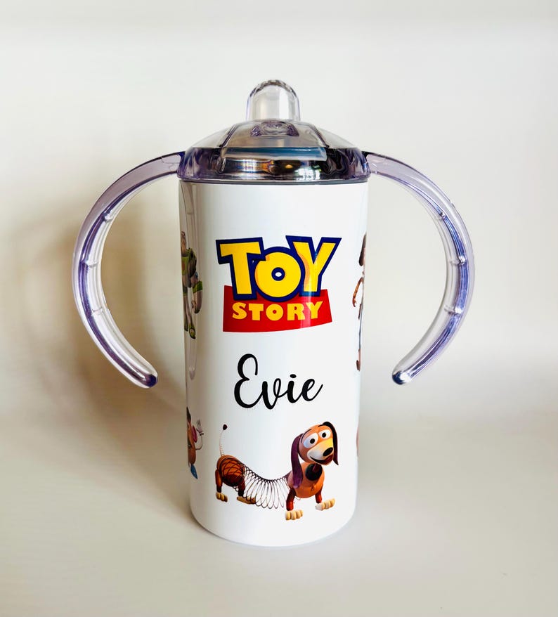 May include: White sippy cup with the "Toy Story" logo in yellow and red, and the name "Evie" in black script. The cup has a clear lid, clear handles, and illustrations of characters from the film.