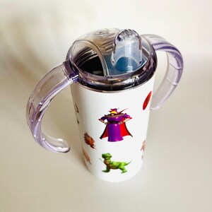 May include: White sippy cup with clear purple handles and a clear lid. The cup features cartoon character graphics, including a purple villain, a green dinosaur, and other colorful images. The cup is designed for toddlers.