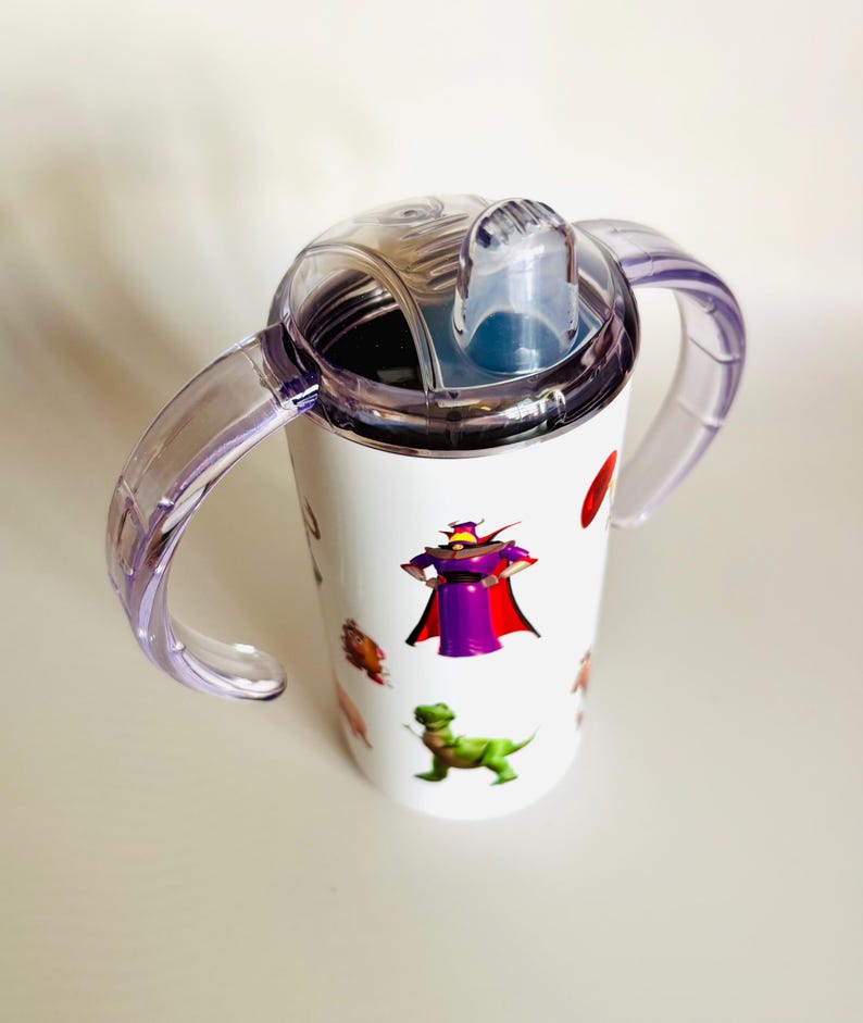 May include: White sippy cup with clear purple handles and a clear lid. The cup features cartoon character graphics, including a purple villain, a green dinosaur, and other colorful images. The cup is designed for toddlers.