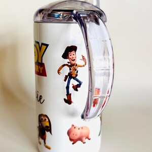 May include: White sippy cup with characters from the Toy Story film. The cup has a clear handle and lid. The design includes Woody, Slinky Dog, Hamm, and the Toy Story logo. The cup is designed for children.