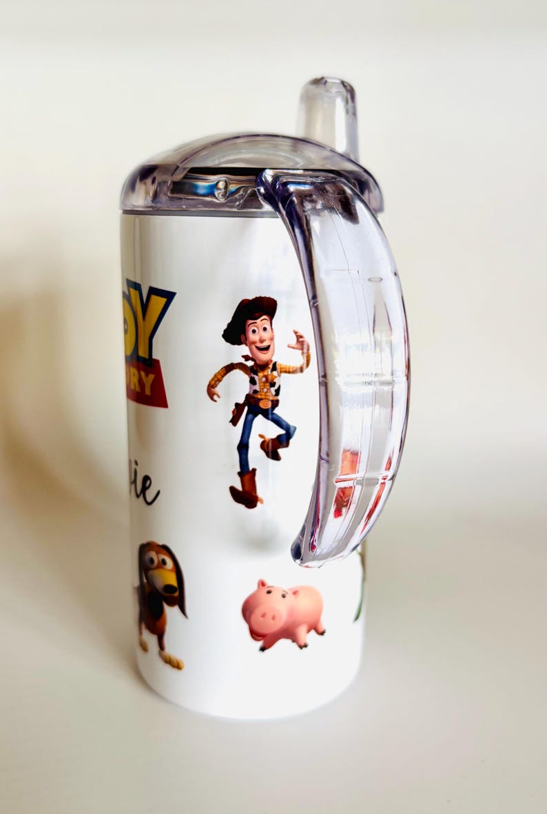 May include: White sippy cup with characters from the Toy Story film. The cup has a clear handle and lid. The design includes Woody, Slinky Dog, Hamm, and the Toy Story logo. The cup is designed for children.