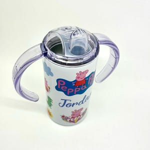 Pode incluir: Um copo de a&ccedil;o inoxid&aacute;vel branco com uma tampa transparente e duas al&ccedil;as roxas. O copo apresenta um design de Peppa Pig com o nome "Jorda" impresso no lado.