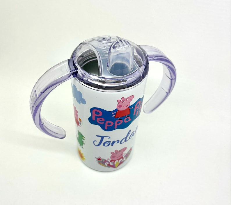 Pode incluir: Um copo de a&ccedil;o inoxid&aacute;vel branco com uma tampa transparente e duas al&ccedil;as roxas. O copo apresenta um design de Peppa Pig com o nome "Jorda" impresso no lado.