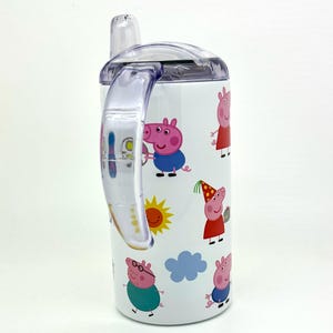 Pode incluir: Um copo de a&ccedil;o inoxid&aacute;vel branco com uma al&ccedil;a e uma tampa de pl&aacute;stico transparente. O copo apresenta um design colorido de personagens do Peppa Pig, incluindo Peppa, George, Papai Pig e Mam&atilde;e Pig. O copo tem um canudo.
