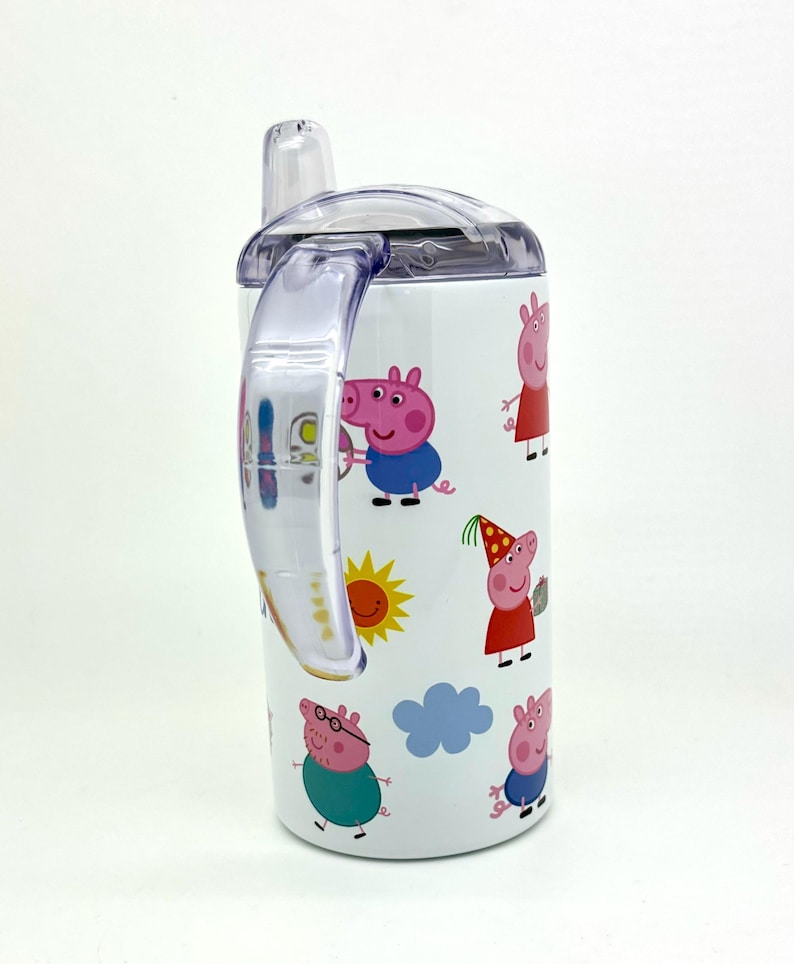 Pode incluir: Um copo de a&ccedil;o inoxid&aacute;vel branco com uma al&ccedil;a e uma tampa de pl&aacute;stico transparente. O copo apresenta um design colorido de personagens do Peppa Pig, incluindo Peppa, George, Papai Pig e Mam&atilde;e Pig. O copo tem um canudo.