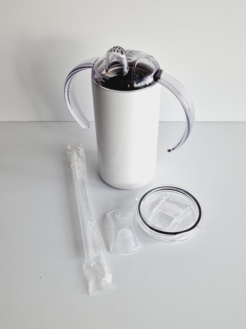 Pode incluir: Um copo de a&ccedil;o inoxid&aacute;vel branco com duas al&ccedil;as, uma tampa transparente com um furo para canudo e um canudo transparente. O copo est&aacute; sobre uma superf&iacute;cie branca.