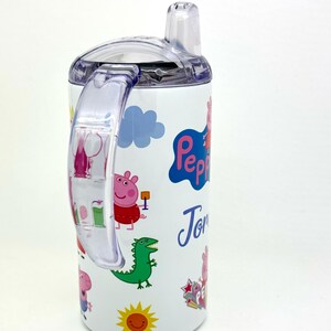 Pode incluir: Garrafa de &aacute;gua de a&ccedil;o inoxid&aacute;vel branca com tampa e al&ccedil;a transparentes. A garrafa apresenta um design de Peppa Pig com um dinossauro verde, um sol e o nome "Tom" escrito no lado.