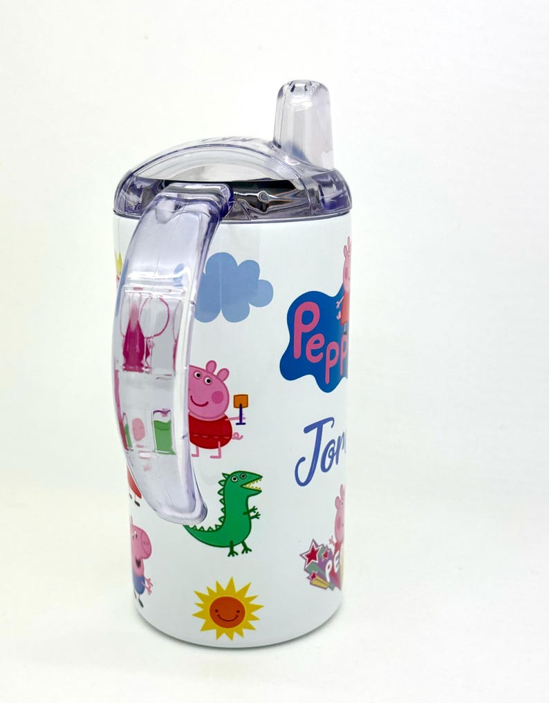 Pode incluir: Garrafa de &aacute;gua de a&ccedil;o inoxid&aacute;vel branca com tampa e al&ccedil;a transparentes. A garrafa apresenta um design de Peppa Pig com um dinossauro verde, um sol e o nome "Tom" escrito no lado.