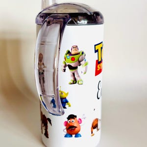 May include: White sippy cup featuring characters from the film Toy Story, including Buzz Lightyear, an alien, and Mr. Potato Head. The cup has a clear handle and lid, and the words "Toy Story" are printed on the side.