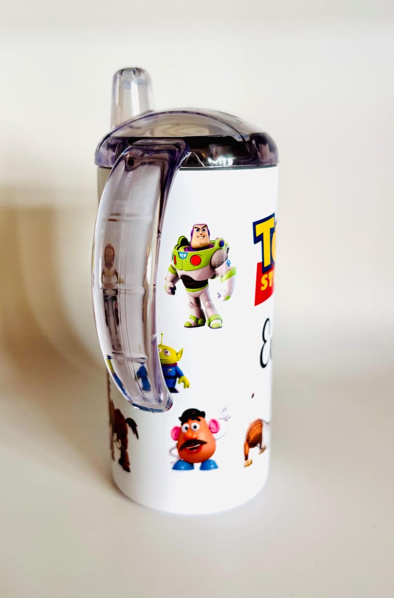May include: White sippy cup featuring characters from the film Toy Story, including Buzz Lightyear, an alien, and Mr. Potato Head. The cup has a clear handle and lid, and the words "Toy Story" are printed on the side.