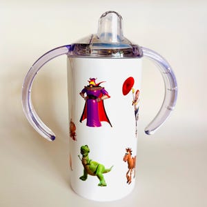 May include: White sippy cup featuring characters from the animated film Toy Story. The cup has a clear lid and handles, with images of Zurg, Rex, and other characters. The cup is designed for toddlers.