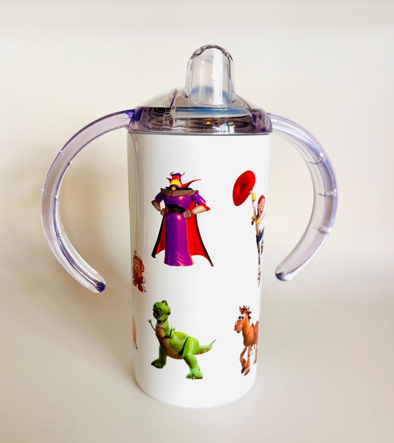 May include: White sippy cup featuring characters from the animated film Toy Story. The cup has a clear lid and handles, with images of Zurg, Rex, and other characters. The cup is designed for toddlers.