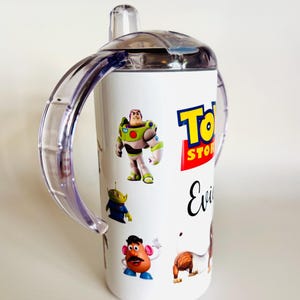 May include: White sippy cup with characters from the Toy Story film, including Buzz Lightyear, an alien, Mr. Potato Head, and Slinky Dog. The cup has a clear lid and handles, and the Toy Story logo is visible.