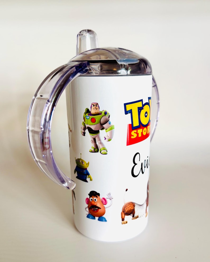 May include: White sippy cup with characters from the Toy Story film, including Buzz Lightyear, an alien, Mr. Potato Head, and Slinky Dog. The cup has a clear lid and handles, and the Toy Story logo is visible.