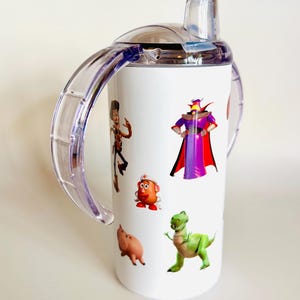 May include: White sippy cup with cartoon character illustrations. The cup has a clear lid and handles, and is decorated with images of characters from the Toy Story film. The cup is designed for toddlers and young children.