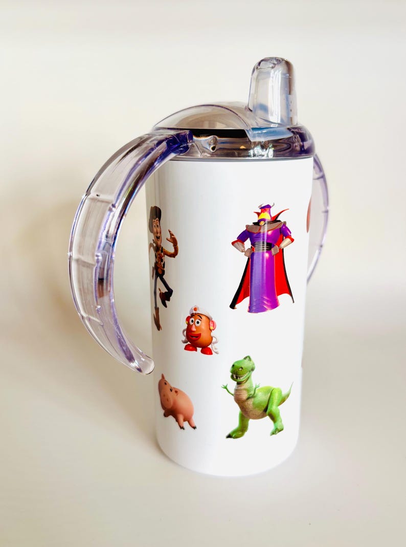 May include: White sippy cup with cartoon character illustrations. The cup has a clear lid and handles, and is decorated with images of characters from the Toy Story film. The cup is designed for toddlers and young children.