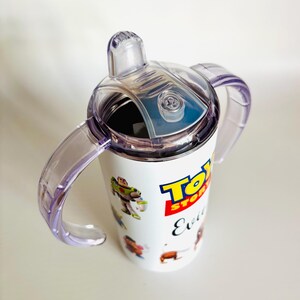 May include: White sippy cup with a clear lid and two purple handles, featuring characters from Toy Story. The Toy Story logo and the word "Evil" are printed on the cup.