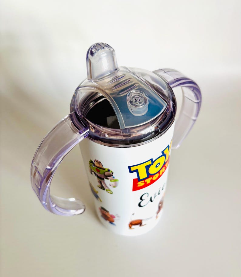 May include: White sippy cup with a clear lid and two purple handles, featuring characters from Toy Story. The Toy Story logo and the word "Evil" are printed on the cup.