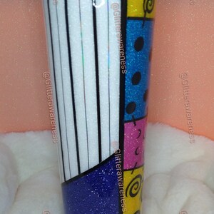 May include: A tall, insulated tumbler with a colorful, patchwork design. The tumbler features a mix of white and black stripes, blue glitter, and sections of yellow, pink, and blue with black accents. A straw is inserted in the lid.
