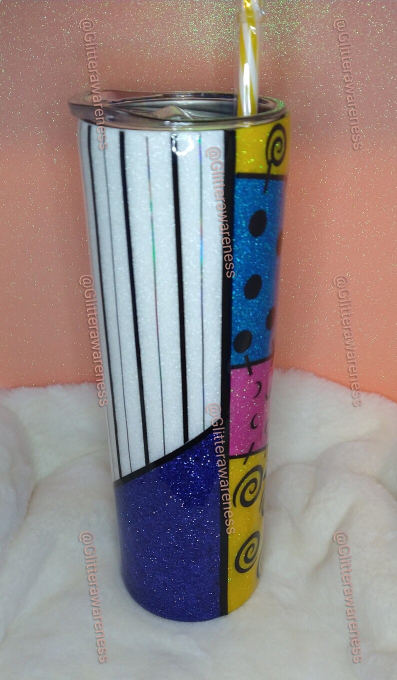 May include: A tall, insulated tumbler with a colorful, patchwork design. The tumbler features a mix of white and black stripes, blue glitter, and sections of yellow, pink, and blue with black accents. A straw is inserted in the lid.
