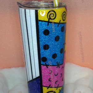 May include: A glitter-covered tumbler with a colorful patchwork design. The tumbler features sections of blue, yellow, pink, and white, with black accents and a striped straw. The design is reminiscent of a character's outfit.