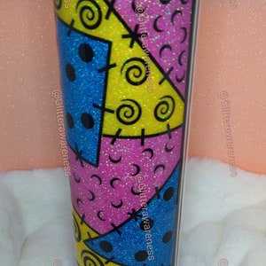 May include: A glitter-covered tumbler with a colorful patchwork design in blue, yellow, and pink. The design includes black stitches, spirals, and dots. A yellow and white striped straw is inserted into the lid.
