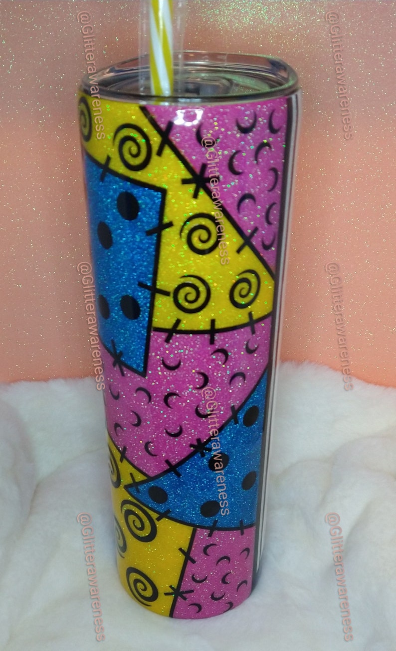 May include: A glitter-covered tumbler with a colorful patchwork design in blue, yellow, and pink. The design includes black stitches, spirals, and dots. A yellow and white striped straw is inserted into the lid.
