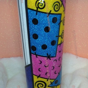 May include: A glitter-covered tumbler with a colorful patchwork design. The design features blue, pink, and yellow sections with black swirls, dots, and cross-stitch details. A yellow and white striped straw is visible.