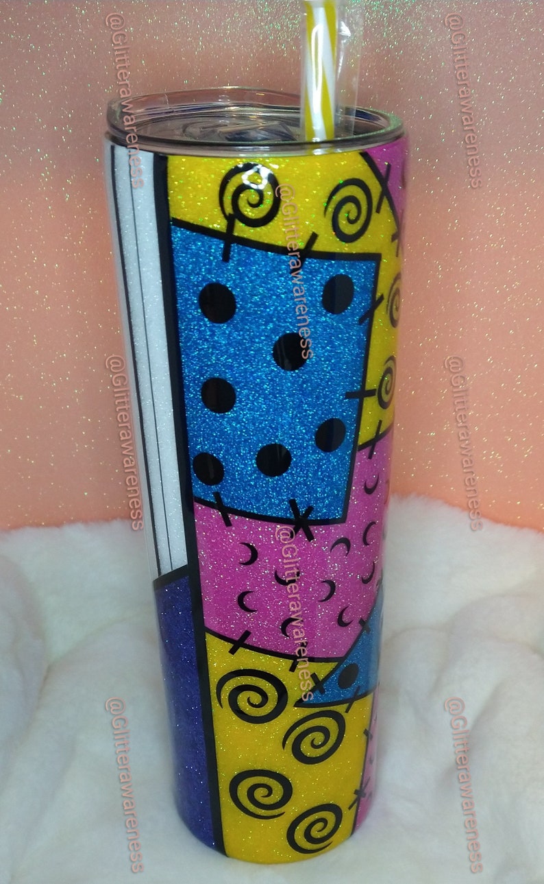 May include: A glitter-covered tumbler with a colorful patchwork design. The design features blue, pink, and yellow sections with black swirls, dots, and cross-stitch details. A yellow and white striped straw is visible.