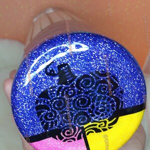May include: A glittery, round bottle stopper with a blue background and a black pumpkin design. The bottom section is divided into pink and yellow sections. The stopper is clear.