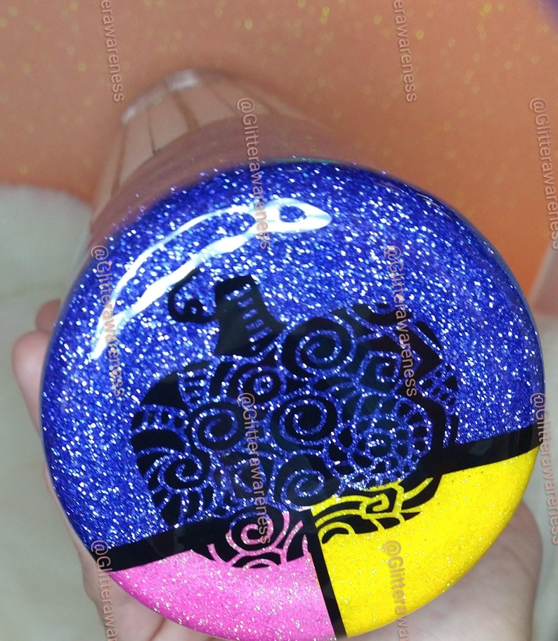 May include: A glittery, round bottle stopper with a blue background and a black pumpkin design. The bottom section is divided into pink and yellow sections. The stopper is clear.